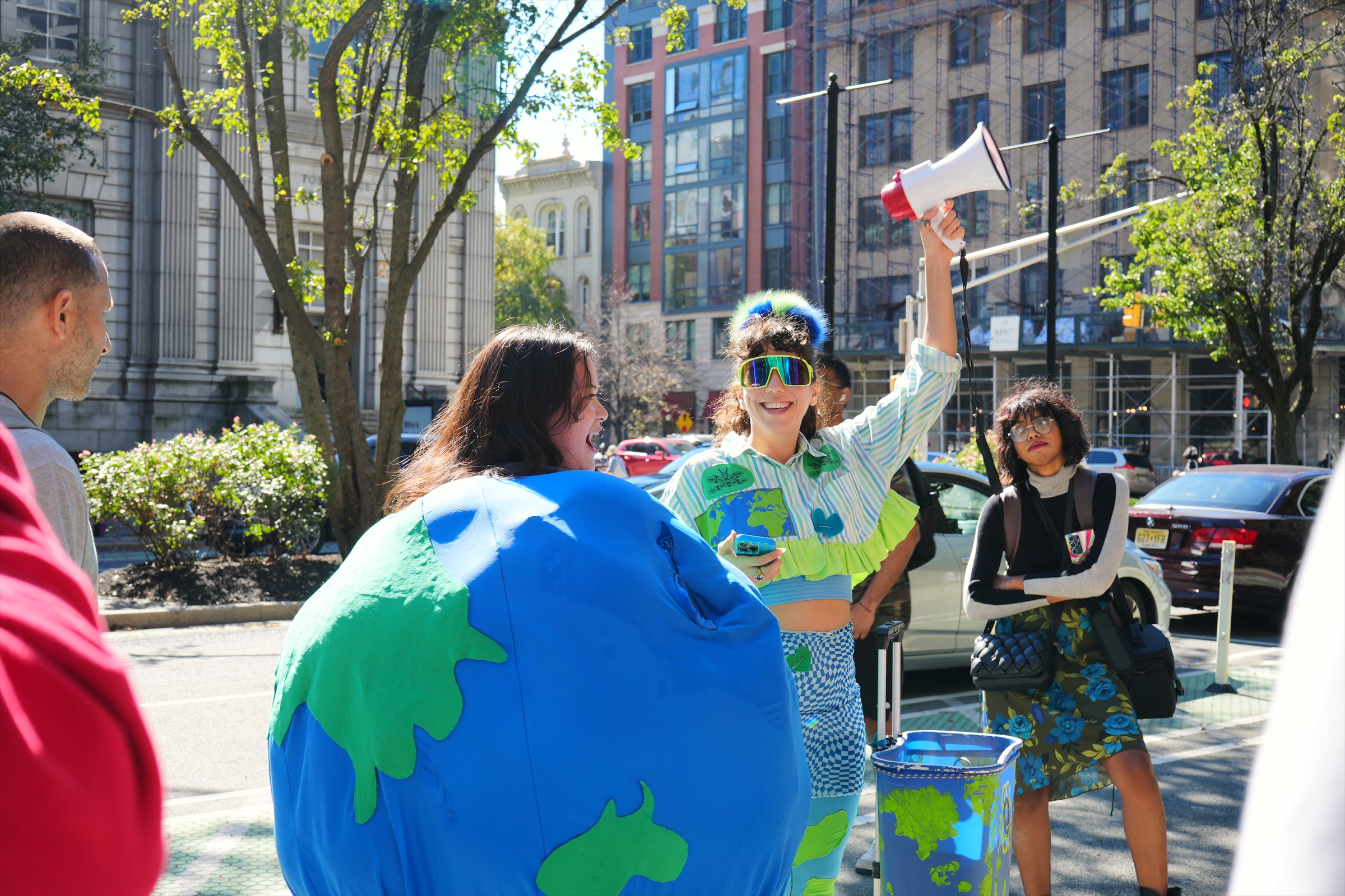 A smiling participant in a striped shirt and colorful Earth-themed costume raises a white megaphone while another marcher stands nearby.