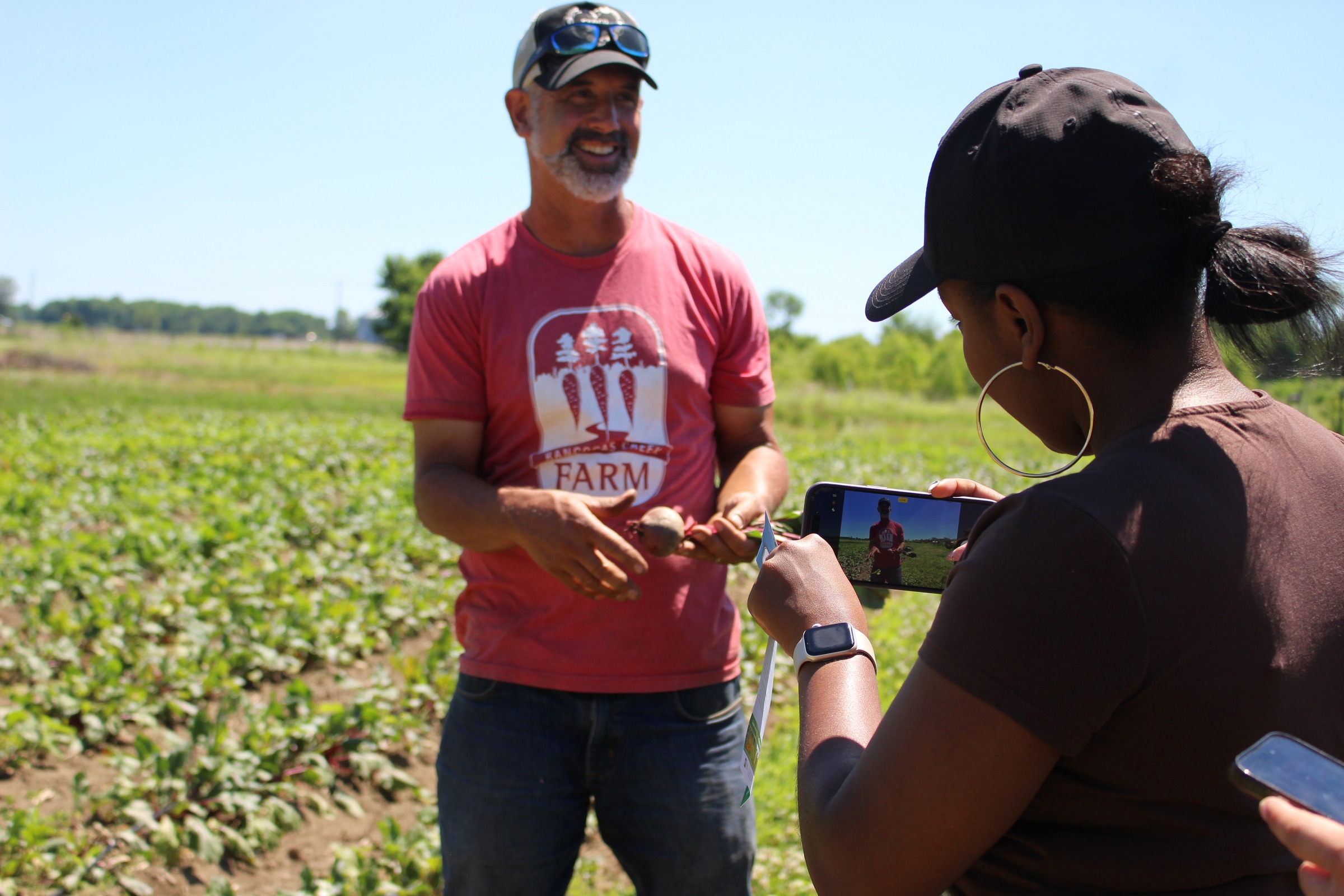A smiling person in a red farm shirt holds a beet while another person records with a phone.