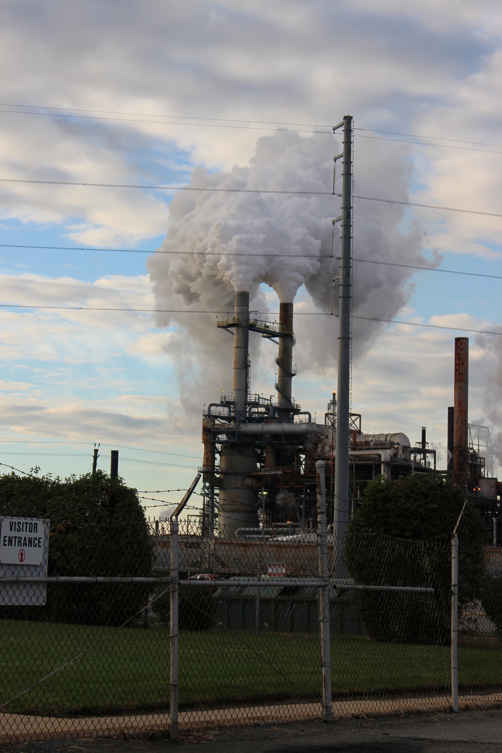 An industrial facility with two tall stacks and pale plumes behind utility wires and a fence.