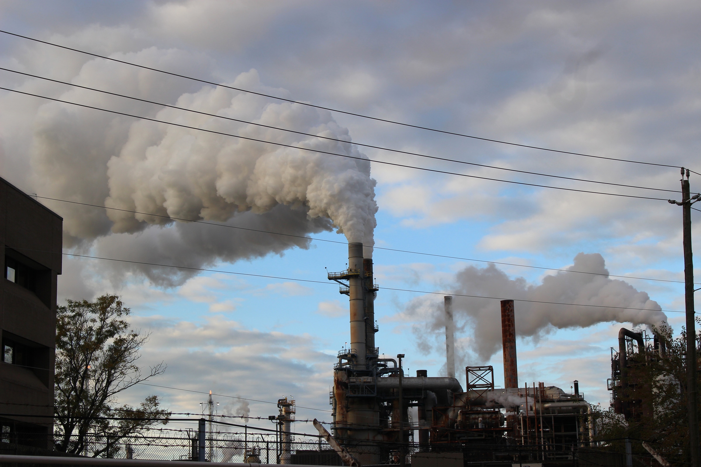 A wide view of industrial structures and pale plumes beneath a cloudy sky, with wires crossing the frame.