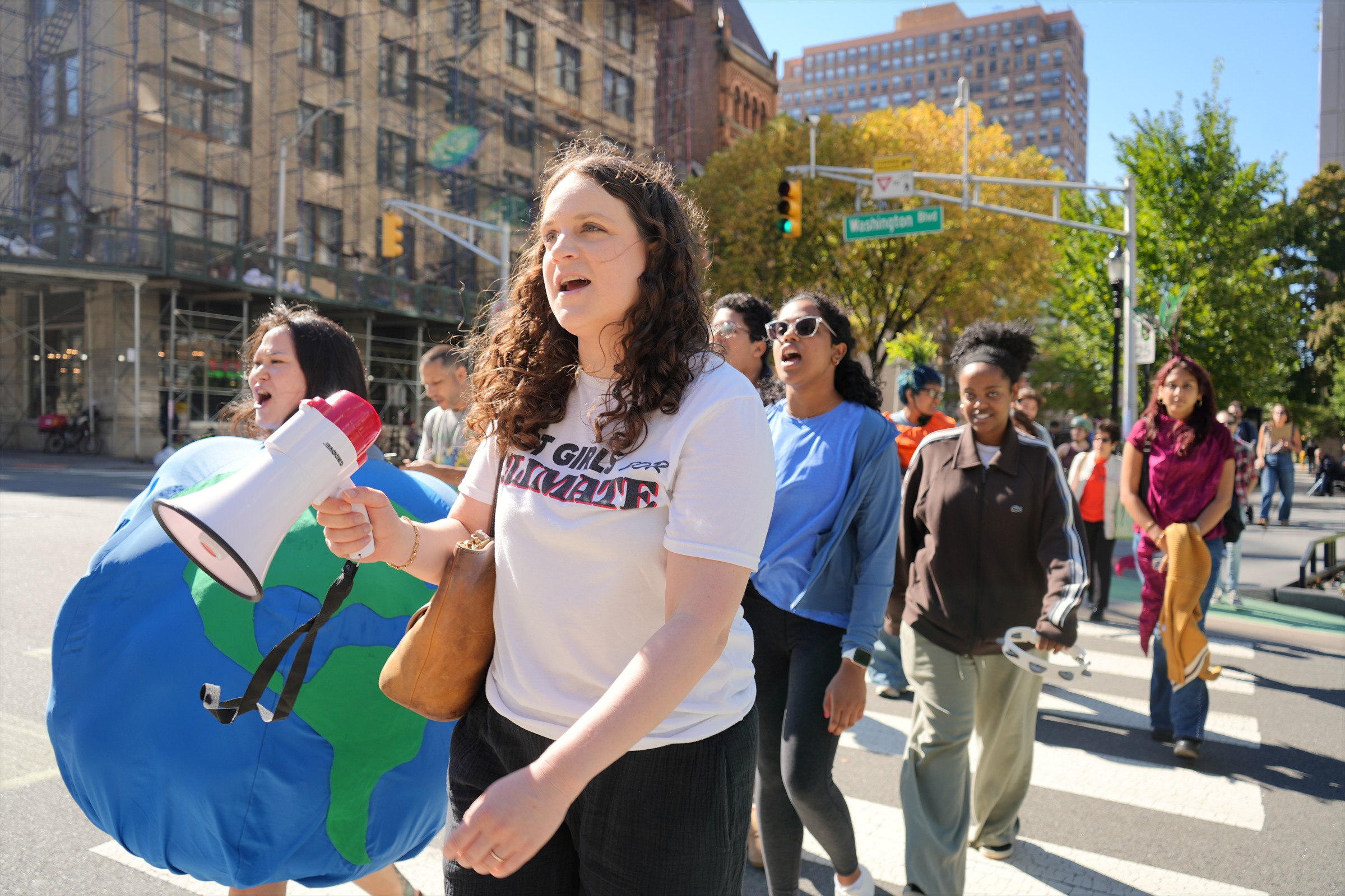 A participant carries a white megaphone as marchers, including a person in a blue-and-green Earth costume, walk along a Jersey City street.