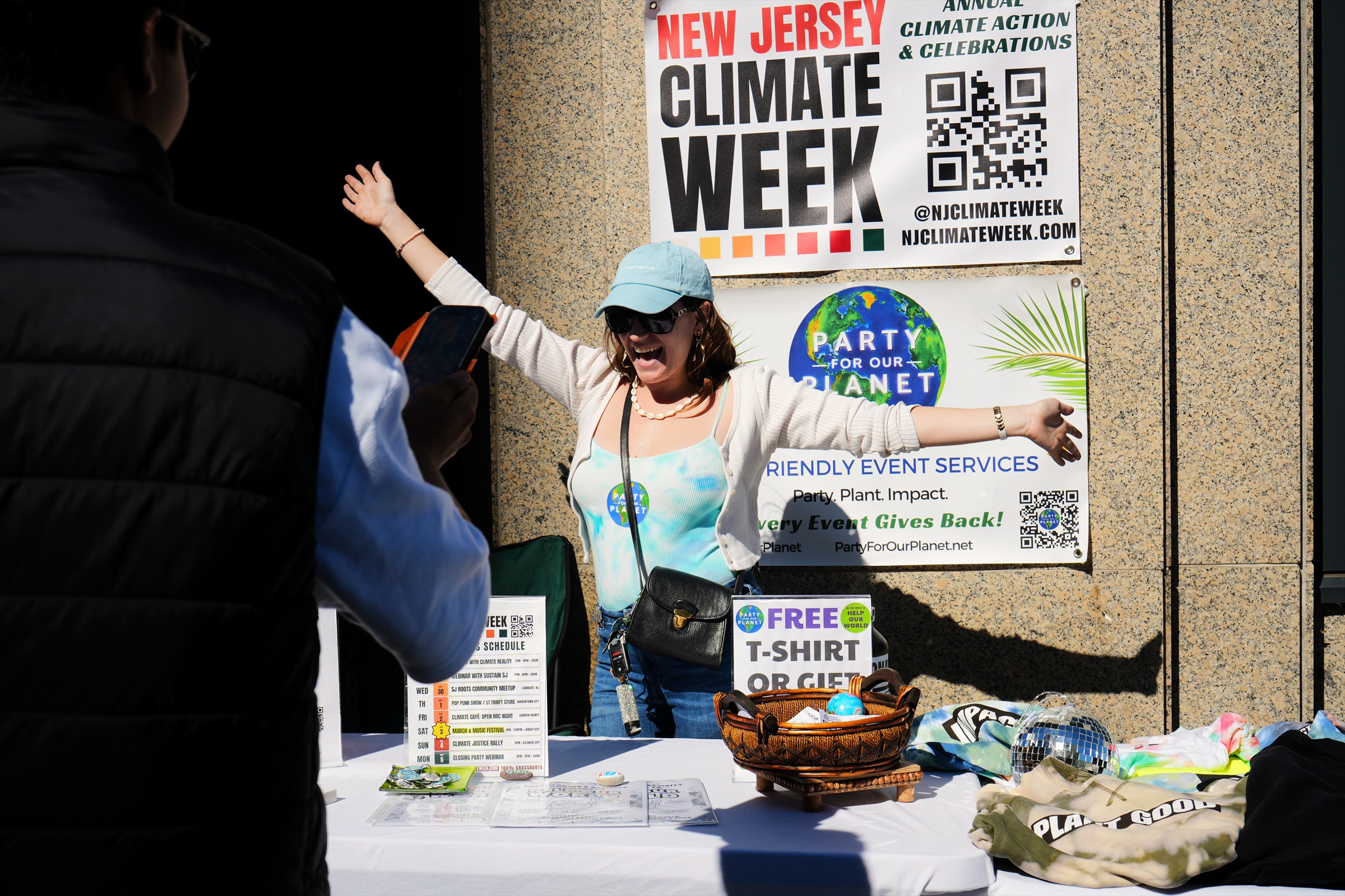 A smiling participant opens both arms beside Party for Our Planet and NJ Climate Week signs at a community table.