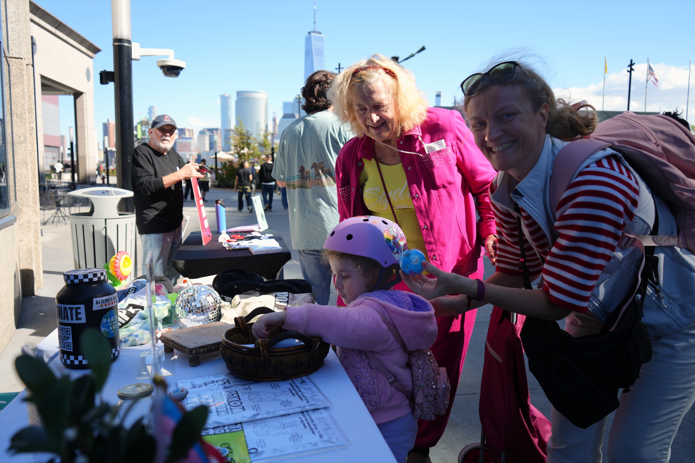 An adult smiles beside a child reaching into a basket at the community table, with another adult in pink standing behind.